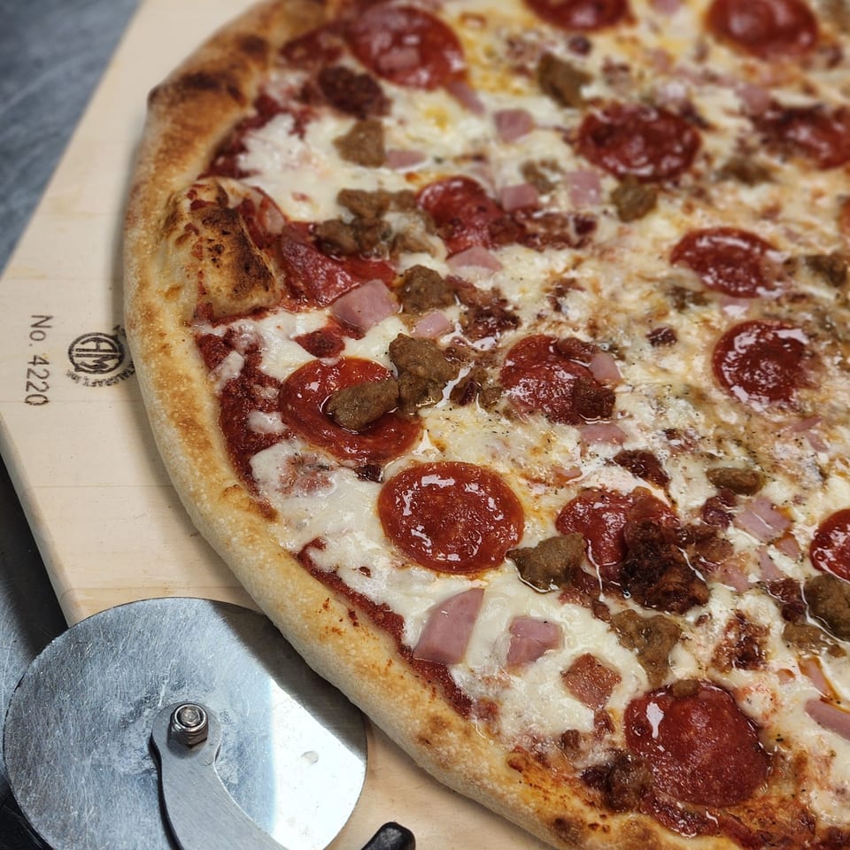 A Slices New York meat lover's pizza with pepperoni, sausage, and ham, on a wooden peel with a cutter.