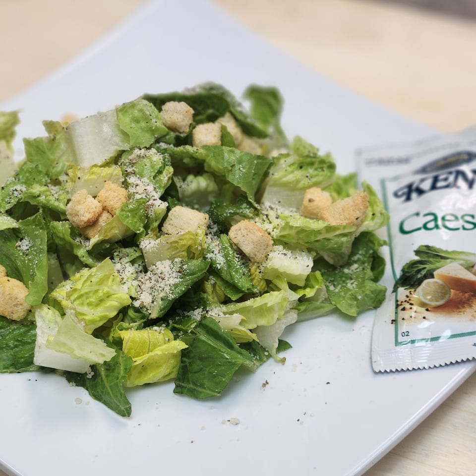 A Caesar salad with croutons and grated cheese on a white plate, with a dressing packet.