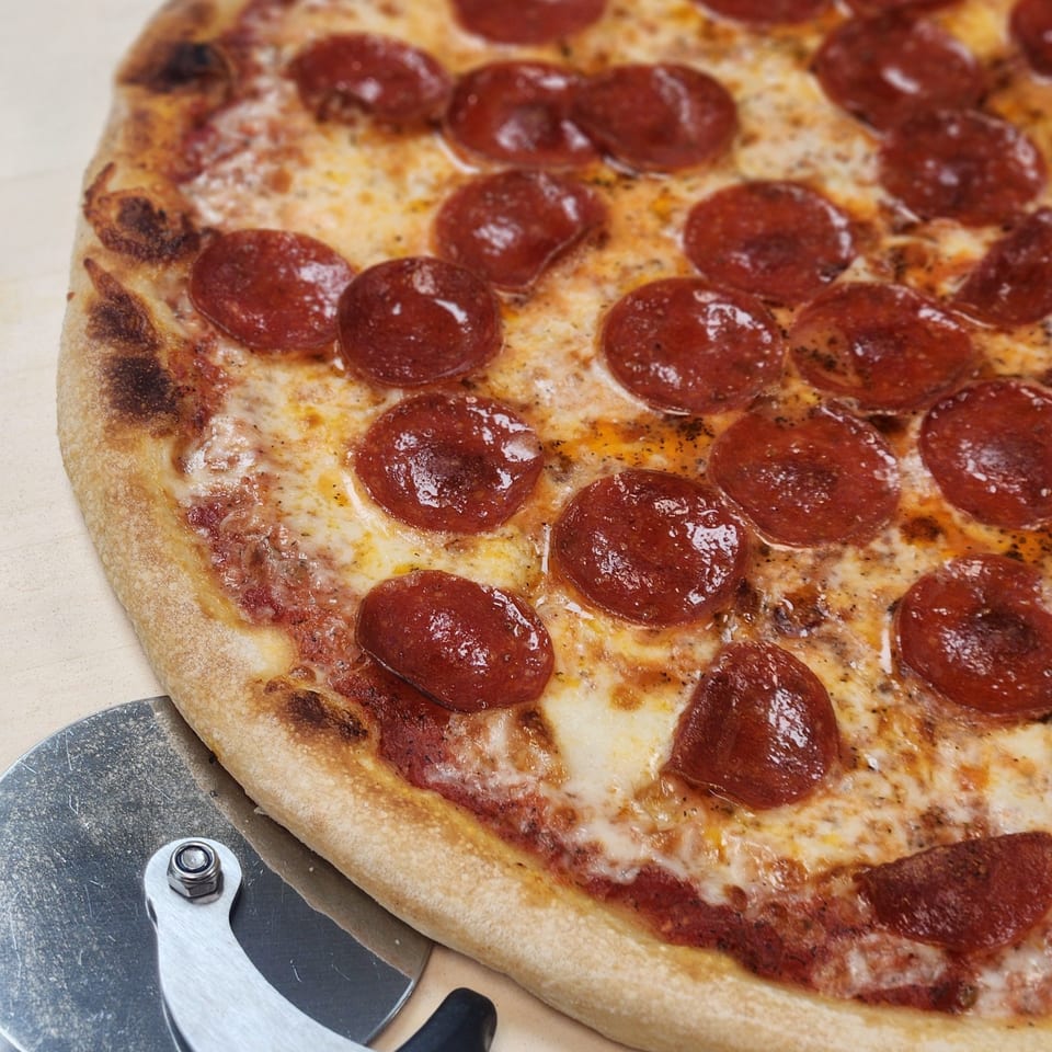 A Slices New York pepperoni pizza with melted cheese and a pizza cutter on a light surface.