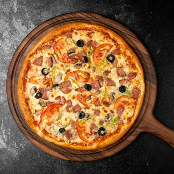 Slices New York pizza with sausage, tomatoes, olives, and mushrooms on a wooden board.