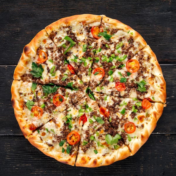 Slices New York pizza with ground meat, melted cheese, cherry tomatoes, and green herbs on a dark wooden table.
